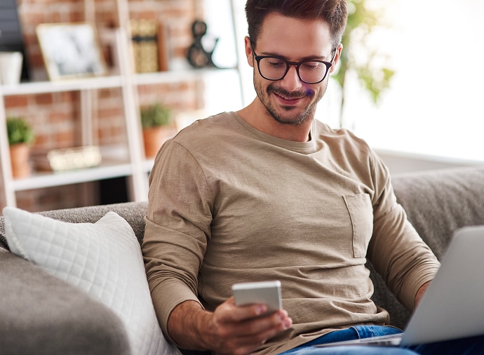 Man sitting on couch looking at his phone while having a laptop on his lap